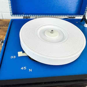 May include: A vintage blue record player with a white turntable. The player has a black front panel and a blue lid with a patterned trim. The speed settings 33 and 45 are visible.