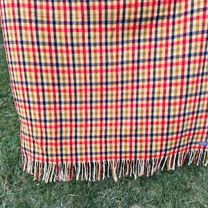 May include: A rectangular woven blanket featuring a multi-colored plaid pattern in shades of red, mustard yellow, and dark navy blue. The fabric has a visible grid design and is finished with a fringe of loose threads along the bottom edge.