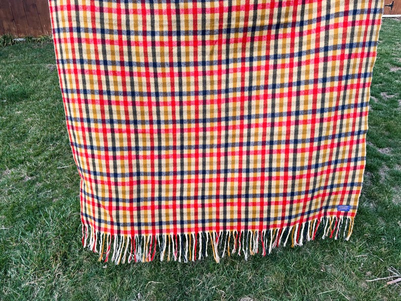 May include: A rectangular woven blanket featuring a multi-colored plaid pattern in shades of red, mustard yellow, and dark navy blue. The fabric has a visible grid design and is finished with a fringe of loose threads along the bottom edge.