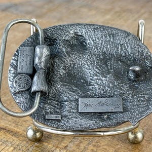 May include: A silver-toned metal belt buckle with a textured oval design. The buckle has a loop, a small rectangular plaque with text, and a decorative element. The back of the buckle is displayed on a metal stand.