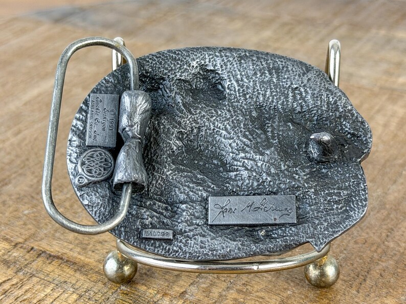 May include: A silver-toned metal belt buckle with a textured oval design. The buckle has a loop, a small rectangular plaque with text, and a decorative element. The back of the buckle is displayed on a metal stand.