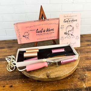May include: Vintage pink Curl-a-Wave electric roller curler set. The set includes a pink box, instructions, curlers, and a curling iron. The box reads "Curl-a-Wave Electric Roller Curler" with a woman's silhouette.