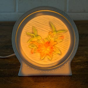 May include: A white ceramic night light with a floral design. The lamp features a circular face with a textured background and a raised relief of yellow and orange flowers. The lamp is lit from within, creating a warm glow.