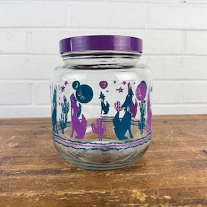 May include: A clear glass jar with a purple lid. The jar features a desert-themed design with teal cacti, purple howling wolves, and stars. The jar sits on a wooden surface against a white brick wall.