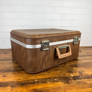 May include: A rectangular brown suitcase with a textured finish, featuring a silver-toned metal trim, two metallic locking latches, and a sturdy plastic handle. The case has a structured design with rounded corners and a flat top surface.