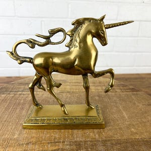 Puede incluir: Una figurita de unicornio de lat&oacute;n dorado con una melena y cola fluidas. El unicornio est&aacute; de pie sobre una base rectangular con un borde decorativo.
