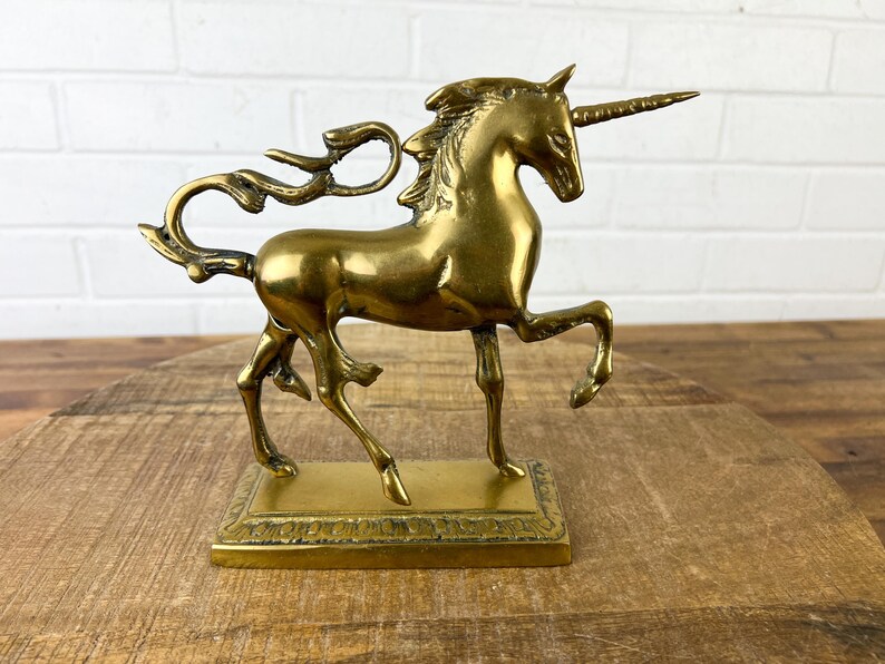 Puede incluir: Una figurita de unicornio de lat&oacute;n dorado con una melena y cola fluidas. El unicornio est&aacute; de pie sobre una base rectangular con un borde decorativo.