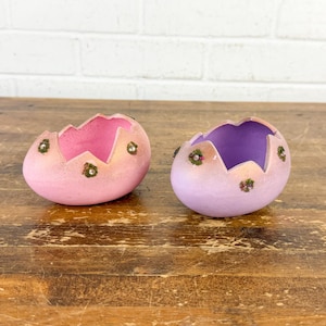 May include: Two decorative, egg-shaped containers. One is light pink with a pink interior, the other is light purple with a purple interior. Both have a jagged edge and small floral accents. They sit on a wooden surface.
