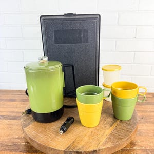 May include: A vintage green and black electric coffee pot with a matching black lunchbox, two sets of stacked yellow and green mugs, and a small container with a yellow lid. The items are arranged on a wooden serving tray.