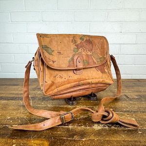 May include: A tan shoulder bag featuring a vintage-style world map print across the exterior. The bag has a fold-over flap with a central snap closure, a long adjustable shoulder strap with a metal buckle, and visible stitching along the edges.