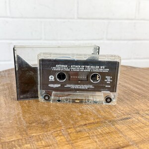May include: A clear cassette tape with the album title "Attack of the Killer B's" by Anthrax. The cassette is in a clear plastic case, resting on a wooden surface. The album title and track list are visible on the label.