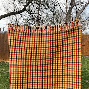 May include: A rectangular woven throw blanket featuring a vibrant plaid pattern in shades of mustard yellow, bright red, and dark navy blue. The blanket is finished with a decorative fringe along the top edge, consisting of alternating colored yarn strands.