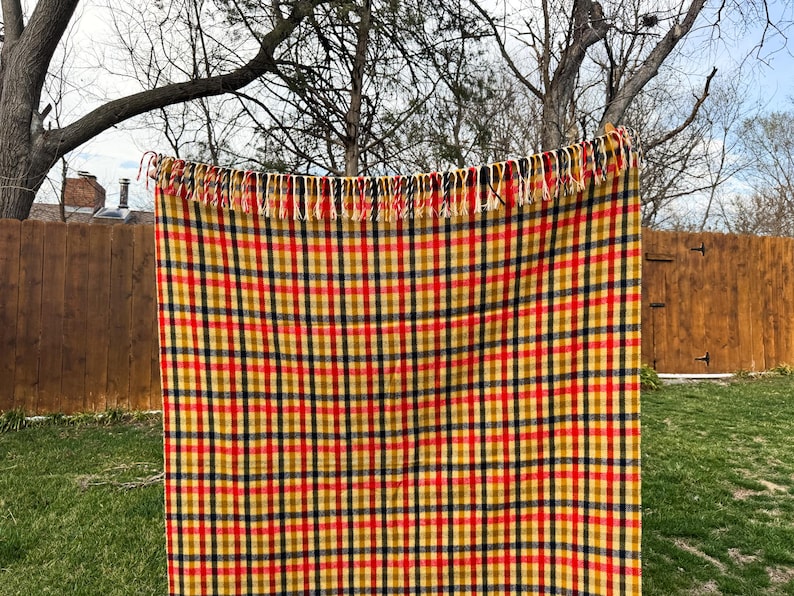 May include: A rectangular woven throw blanket featuring a vibrant plaid pattern in shades of mustard yellow, bright red, and dark navy blue. The blanket is finished with a decorative fringe along the top edge, consisting of alternating colored yarn strands.