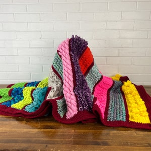 May include: A colorful crocheted blanket with stripes of pink, red, green, blue, yellow, and gray. The blanket is made with different crochet stitches, including bobble stitch, cable stitch, and single crochet.