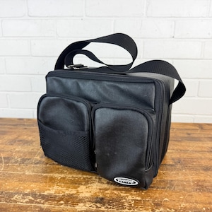 May include: A black rectangular bag with an adjustable shoulder strap, featuring two front zippered pockets, one mesh side pocket, and a small white oval patch with the word CONDUCTOR printed in black text on the lower right corner.