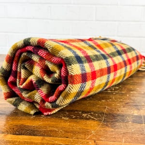 May include: A rolled-up woven blanket featuring a multi-colored plaid pattern in shades of mustard yellow, bright red, dark navy blue, and forest green. The fabric has a visible textured weave with frayed edges visible at the end of the roll.