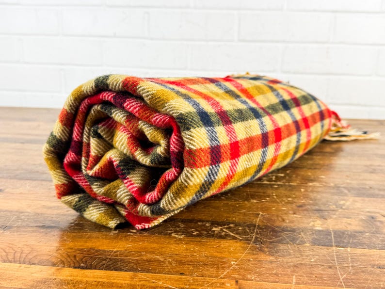 May include: A rolled-up woven blanket featuring a multi-colored plaid pattern in shades of mustard yellow, bright red, dark navy blue, and forest green. The fabric has a visible textured weave with frayed edges visible at the end of the roll.