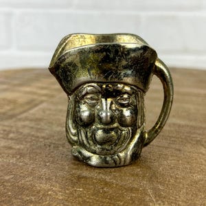 May include: A silver-toned metal pitcher shaped like a pirate's head wearing a hat. The pitcher has a handle and a spout.