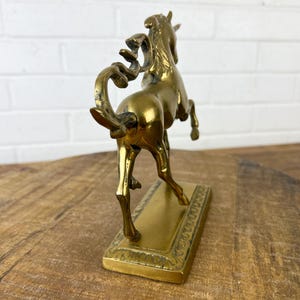 Puede incluir: Una escultura de bronce dorado de un caballo encabritado sobre una base rectangular. El caballo est&aacute; detallado con una melena y una cola fluidas.