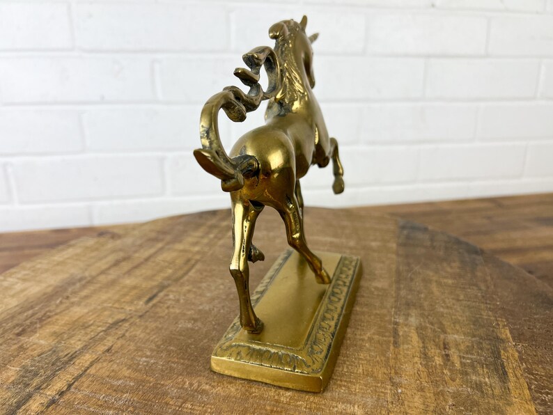 Puede incluir: Una escultura de bronce dorado de un caballo encabritado sobre una base rectangular. El caballo est&aacute; detallado con una melena y una cola fluidas.