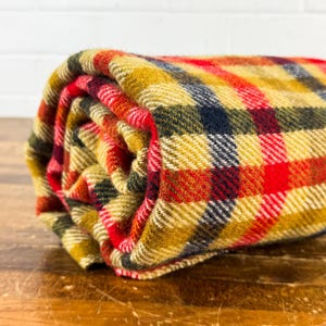 May include: A rolled wool blanket featuring a multi-colored plaid pattern with intersecting lines in shades of mustard yellow, vibrant red, dark navy blue, and forest green. The fabric has a visible woven texture throughout the entire surface.