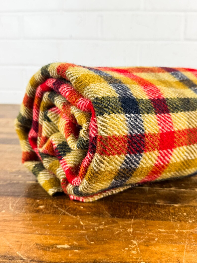 May include: A rolled wool blanket featuring a multi-colored plaid pattern with intersecting lines in shades of mustard yellow, vibrant red, dark navy blue, and forest green. The fabric has a visible woven texture throughout the entire surface.