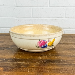 May include: Cream-colored ceramic mixing bowl with a painted fruit design. The bowl features a light tan rim and a decorative band with a pink apple, blue grapes, and a yellow pear. The bowl sits on a wooden surface.