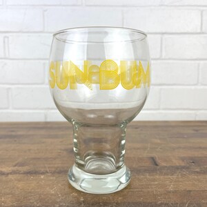 May include: Clear glass beer glass with a stem and a wide bowl. The words "SUN & BUM" are printed in yellow block letters on the glass. The glass is sitting on a wooden surface.