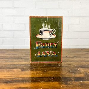 May include: A green wooden sign with a red border and the words "Fancy Java" in colorful letters. A white coffee cup with steam rising from it is pictured above the text.