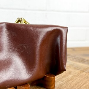 May include: A brown leather coin purse with a brass clasp. The purse is rectangular with rounded edges and a slightly textured surface. The clasp is a simple, hinged design.