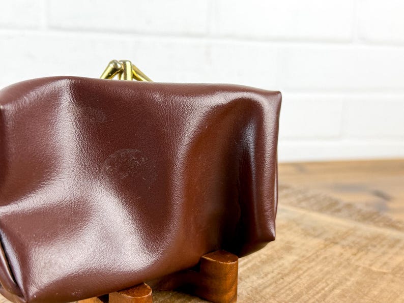 May include: A brown leather coin purse with a brass clasp. The purse is rectangular with rounded edges and a slightly textured surface. The clasp is a simple, hinged design.