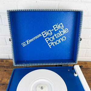 May include: A vintage Emerson Big-Big Portable Phono record player. The blue vinyl case is open, revealing a white turntable and the text "Emerson Big-Big Portable Phono" in white. The case is trimmed with a blue and white patterned fabric.