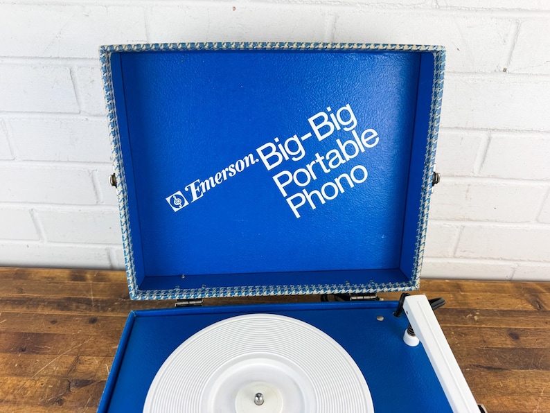 May include: A vintage Emerson Big-Big Portable Phono record player. The blue vinyl case is open, revealing a white turntable and the text "Emerson Big-Big Portable Phono" in white. The case is trimmed with a blue and white patterned fabric.