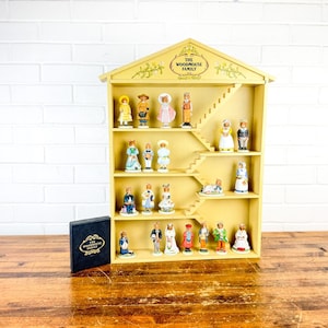 May include: A yellow wooden shelf unit shaped like a house, filled with small figurines. The shelf has a staircase design and the text "THE WOODHOUSE FAMILY" at the top. A black box with gold text sits in front of the shelf.