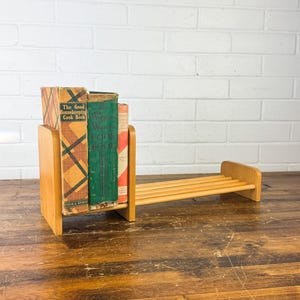 May include: A wooden bookshelf with three vintage cookbooks. The books have colorful covers with titles like "The Good Housekeeping Cook Book" and "American Woman's Cook Book". The shelf has a built-in rack.