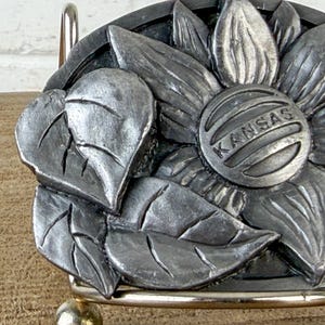 May include: A silver-toned metal belt buckle featuring a detailed sunflower design with leaves. The center of the flower has the word "KANSAS" engraved. The buckle is mounted on a silver-coloured metal frame.