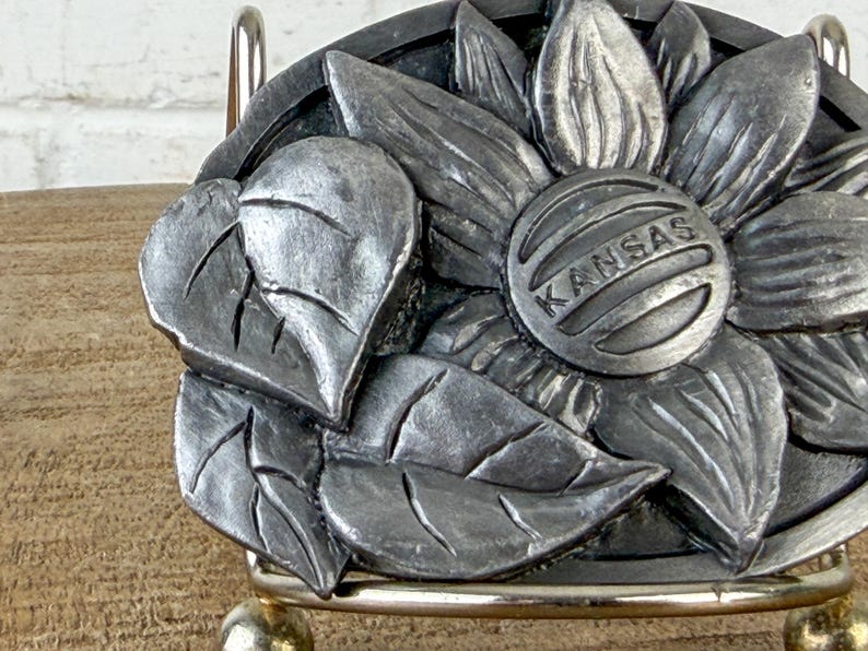 May include: A silver-toned metal belt buckle featuring a detailed sunflower design with leaves. The center of the flower has the word "KANSAS" engraved. The buckle is mounted on a silver-coloured metal frame.