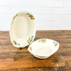 May include: Two off-white oval serving bowls with scalloped edges and floral accents in red, blue, and green. The bowls are on a wooden surface, with a white brick wall in the background. The larger bowl is slightly elevated.