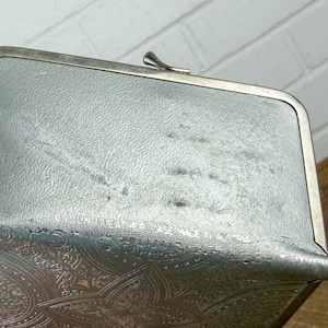 May include: A vintage silver-toned coin purse with a decorative embossed pattern on the lower portion. The purse has a clasp closure and a textured surface. The words "Genuine Leather" are visible on the top of the purse.