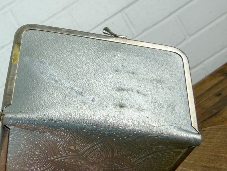 May include: A vintage silver-toned coin purse with a decorative embossed pattern on the lower portion. The purse has a clasp closure and a textured surface. The words "Genuine Leather" are visible on the top of the purse.