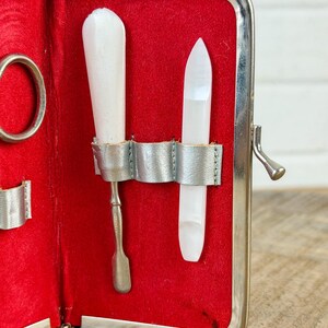 May include: Open red manicure kit with silver trim. The kit contains a white nail buffer, a silver cuticle pusher, and a white nail file. The kit is open, revealing the tools against a red velvet interior.