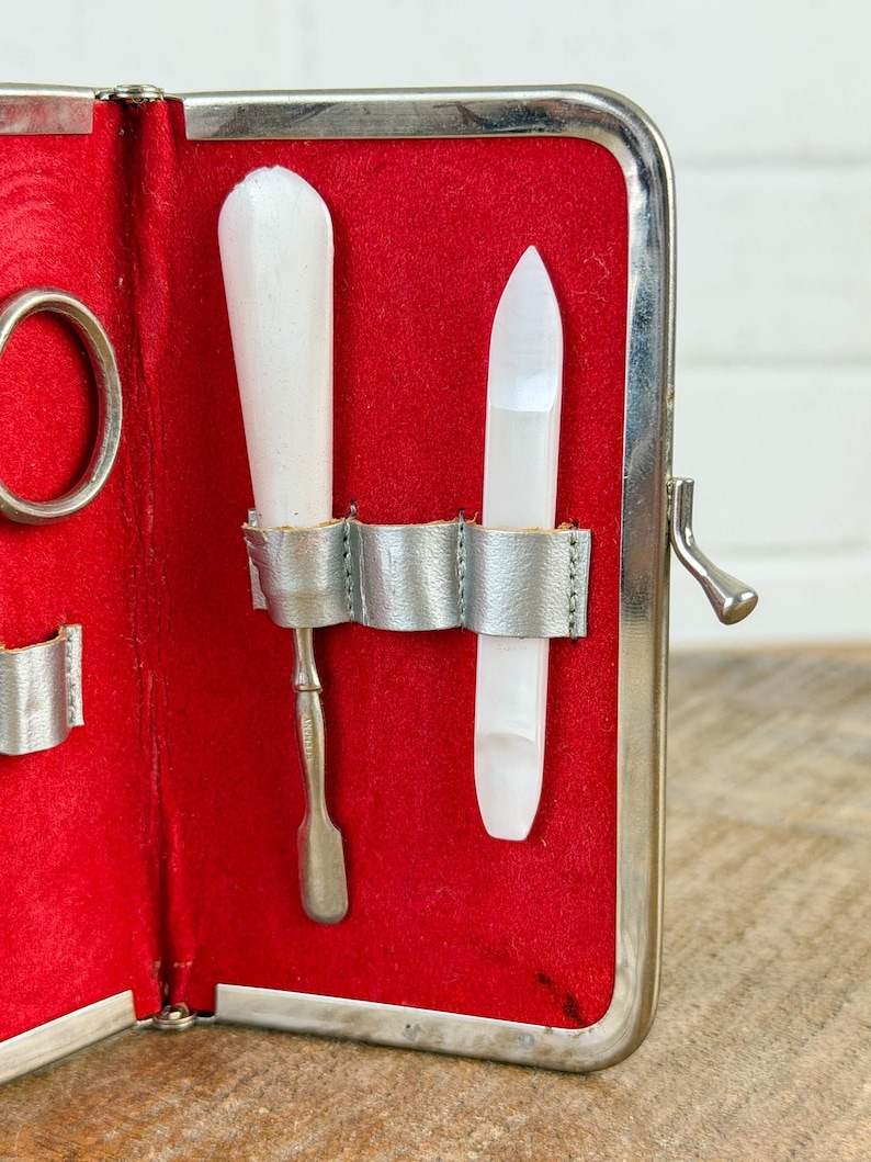 May include: Open red manicure kit with silver trim. The kit contains a white nail buffer, a silver cuticle pusher, and a white nail file. The kit is open, revealing the tools against a red velvet interior.