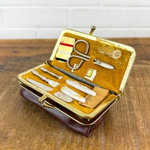 May include: A vintage brown leatherette sewing kit with a gold-toned clasp, open to reveal various sewing tools. The kit includes small scissors, needles, buttons, and other accessories, all neatly arranged on a mustard-yellow velvet lining. A classic and compact travel sewing kit.