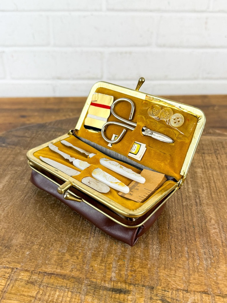 May include: A vintage brown leatherette sewing kit with a gold-toned clasp, open to reveal various sewing tools. The kit includes small scissors, needles, buttons, and other accessories, all neatly arranged on a mustard-yellow velvet lining. A classic and compact travel sewing kit.
