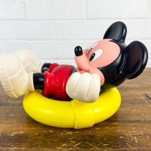 Puede incluir: Una figura vintage de Mickey Mouse en un flotador inflable amarillo. Mickey lleva pantalones cortos rojos, guantes blancos y zapatos color crema. La figura tiene orejas negras y una cara rosa. El juguete est&aacute; sobre una superficie de madera.