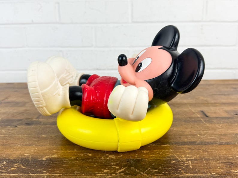 Puede incluir: Una figura vintage de Mickey Mouse en un flotador inflable amarillo. Mickey lleva pantalones cortos rojos, guantes blancos y zapatos color crema. La figura tiene orejas negras y una cara rosa. El juguete est&aacute; sobre una superficie de madera.