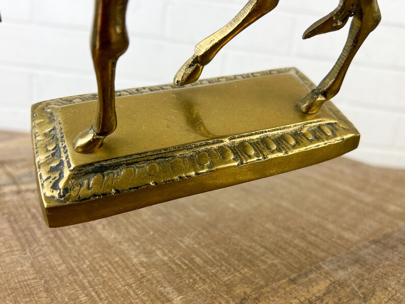 Puede incluir: Una escultura de bronce dorado de un ciervo saltando con una base ornamentada. El ciervo est&aacute; en el aire con las patas extendidas. La base es rectangular con un borde decorativo.