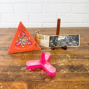 May include: A vintage Pixie Bow Maker set. The set includes a bright pink plastic bow-making tool, a triangular orange box with floral graphics, and an instruction booklet. The box reads "Have fun, make bow after bow the easy way."