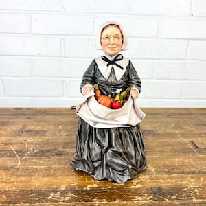 May include: A ceramic figurine of a woman in traditional colonial attire, holding a white sack filled with produce. The figure wears a black dress, white collar, and a white bonnet. The figurine is on a wooden surface.