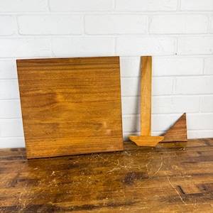 May include: A wooden square, a T-shaped piece, and a triangular piece are arranged on a wooden surface. The wood has a warm, medium brown tone with visible grain. The background is a white brick wall.